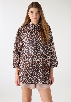 Giovane donna con i capelli lunghi che indossa una camicia a maniche lunghe con stampa leopardata, sciolta e abbottonata, e pantaloncini di colore chiaro su uno sfondo semplice.