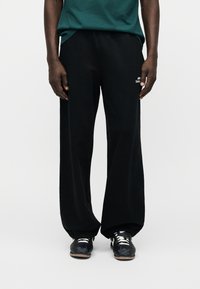 Άνδρας που φοράει φαρδιές μαύρες φόρμες με ελαστικά τελειώματα και μαύρα αθλητικά παπούτσια με λευκά κορδόνια, στέκεται μπροστά σε λευκό φόντο.