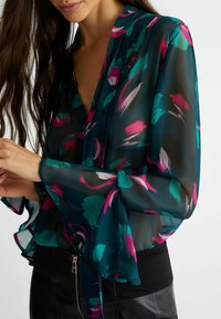 Blusa trasparente e stampata con maniche lunghe a campana. Presenta design a pennellate in turchese, rosa e bianco su uno sfondo scuro. Scollo a V.