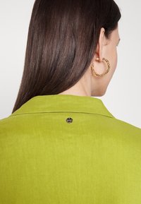 Donna con lunghi capelli castani che indossa una giacca verde lime con un logo metallico sul colletto e orecchini a cerchio dorati attorcigliati, di spalle.
