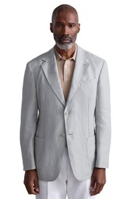 Blazer grigio chiaro in lino con rever a incavo, due tasche frontali e una tasca sul petto. Indossato sopra una polo beige.