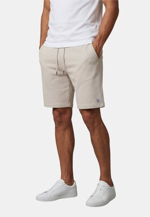 Mann trägt beige Shorts mit Kordelzug, weißes T-Shirt und weiße Sneakers, steht mit den Händen in den Taschen vor einfarbigem Hintergrund.