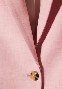 Jasnoróżowy żakiet z teksturowanej tkaniny, z klapami i dużym brązowym guzikiem na lewym przedzie.
