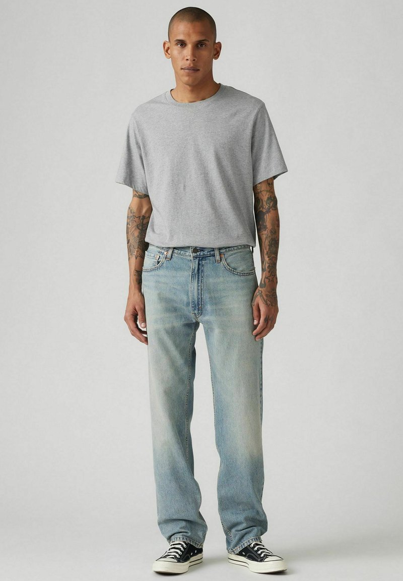 Hombre con tatuajes que lleva una camiseta gris sencilla, jeans claros descoloridos y zapatillas negras de caña baja, de pie contra un fondo liso.