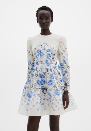 Femeie purtând o rochie albă cu motive florale albastre, mâneci lungi bufante și talie ajustată, stând în fața unui fundal simplu.