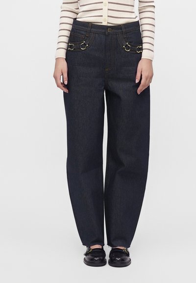 Personne portant un jean bleu foncé à jambes larges avec des anneaux métalliques décoratifs sur les poches avant, un pull léger rayé et des mocassins noirs.