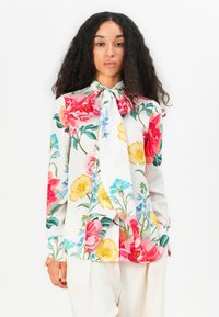 Blusa floreale con maniche lunghe, caratterizzata da uno sfondo bianco, fiori multicolori in rosa, giallo e blu, e un dettaglio del colletto annodato.