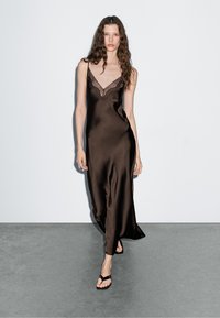 Braunes Satin-Nachtkleid mit Spitzenbesatz am Ausschnitt, dünnen Trägern und fließendem Design. Getragen mit einfachen schwarzen Flip-Flops.