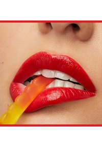 Gros plan de lèvres portant un rouge à lèvres rouge vif mordant un bonbon en forme de ver gélifié bicolore avec des dents visibles et une partie du nez au-dessus.