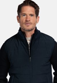 Dunkelblaues gepolstertes Westchen mit hohem Kragen, Reißverschluss vorne und gerippten schwarzen Sweaterärmeln. Mit horizontal gestepptem Muster und Seitentaschen.