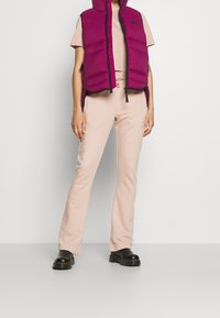 Lila pufferväst med svarta detaljer, bärs över en ljusrosa topp och matchande byxor med en dekorativ detalj. Svarta robusta skor fulländar looken.