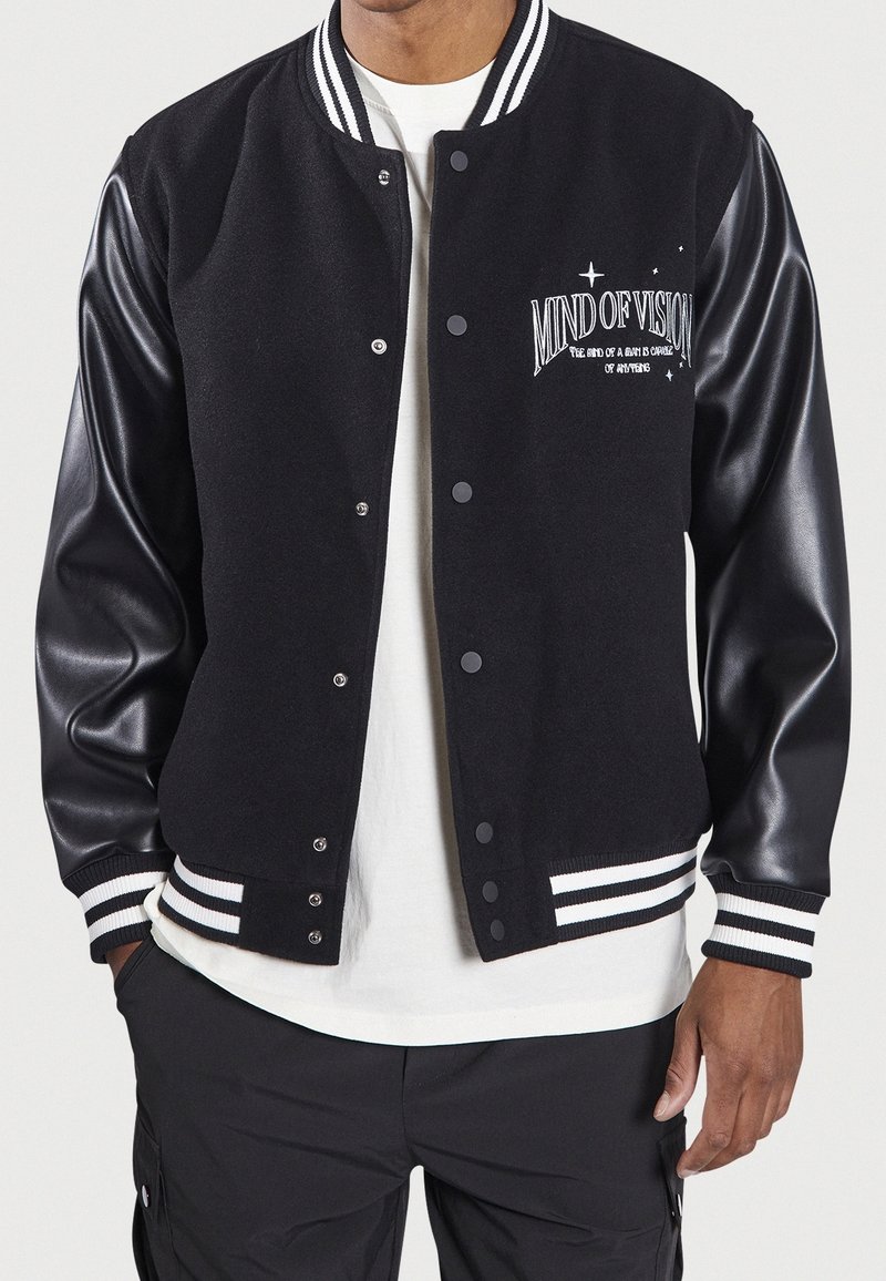 Homme portant une veste varsity noire avec des manches en cuir et un texte brodé blanc "MIND OF VISION" sur la poitrine gauche, associée à une chemise blanche et un pantalon noir.