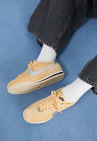 Person med beige Nike-sneakers, vita strumpor och mörka jeans, sittande på en blå heltäckningsmatta.
