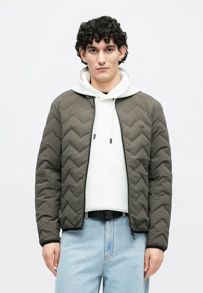 Olivovo zelená prešívaná bunda s chevronovým vzorom, predným zipsom a rebrovanými manžetami, nosená cez bielu mikinu s kapucňou a svetlomodré džínsy.