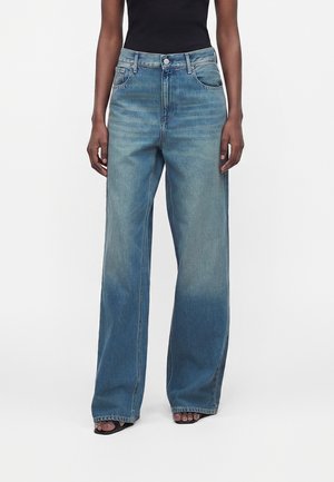 Άτομο που φοράει μπλε τζιν με ψηλή μέση και φαρδιά μπατζάκια με ξεθωριασμένες λεπτομέρειες, σε συνδυασμό με μαύρες ανοιχτές γόβες και μαύρο αμάνικο κορμάκι.