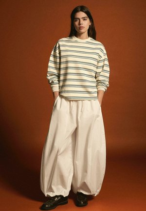 Frau steht mit den Händen in den Taschen, trägt einen beige gestreiften Pullover, breite weiße Hosen und schwarze Schuhe vor braunem Hintergrund.