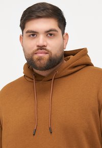 Brązowa bluza z kapturem, wykonana z miękkiego materiału, z regulowanym kapturem z czarnymi końcówkami oraz luźnym krojem. Prosty design bez wzorów.