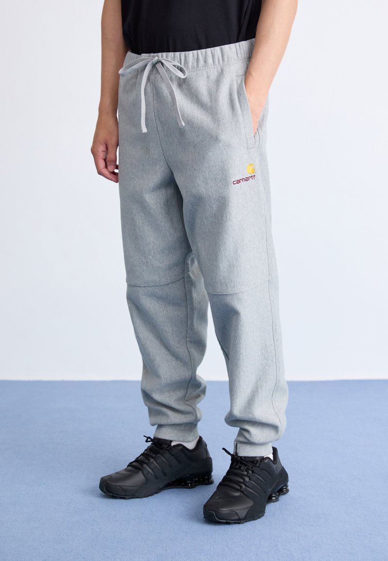 Graue Sweatpants mit elastischem Bund und Kordelzug, ausgestattet mit einem roten Carhartt-Logo auf dem linken Oberschenkel, kombiniert mit schwarzen Sportschuhen.