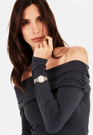 Sølv ur med mesh rem på en kvindes håndled, iført en mørk off-shoulder top. Uret har en struktureret urskive og minimalistisk design.