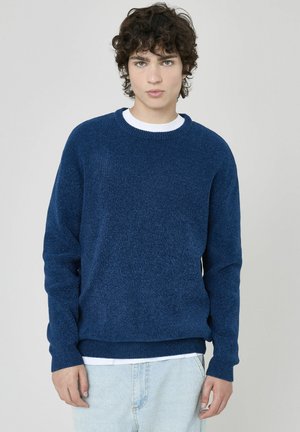 Pull en maille bleu marine avec un col rond classique et des poignets côtelés. Porté par-dessus une chemise blanche superposée, avec une finition texturée.