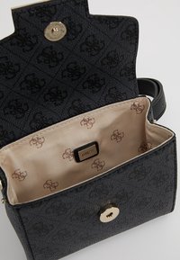 Lille sort struktureret håndtaske med mønstret foring og guldknaplukning, åben for at vise beige indvendig med gentaget logo og "GUESS"-mærke.
