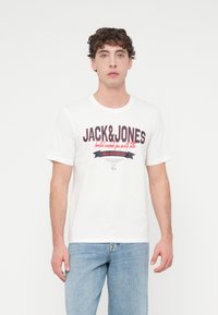 Λευκό βαμβακερό T-shirt με μεγάλο λογότυπο "JACK&JONES" σε μαύρο και κόκκινο, με λεπτομέρεια κορδέλας. Συνδυασμένο με ανοιχτόχρωμα μπλε τζιν παντελόνια.