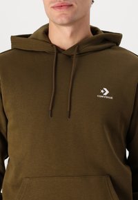 Sudadera verde oliva hecha de una tela suave y ligera. Cuenta con un bolsillo canguro, capucha con cordón y un logo blanco de "Converse".