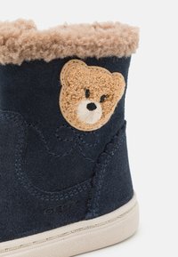 Stivale in suede navy con collare in pelliccia beige, caratterizzato da una toppa a forma di orso testurizzata e dettagli di cucitura sottili. Suola in gomma resistente.