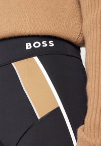 Gros plan sur un legging noir avec des rayures verticales beige et blanches, logo BOSS sur la ceinture, associé à un pull en tricot couleur chameau.