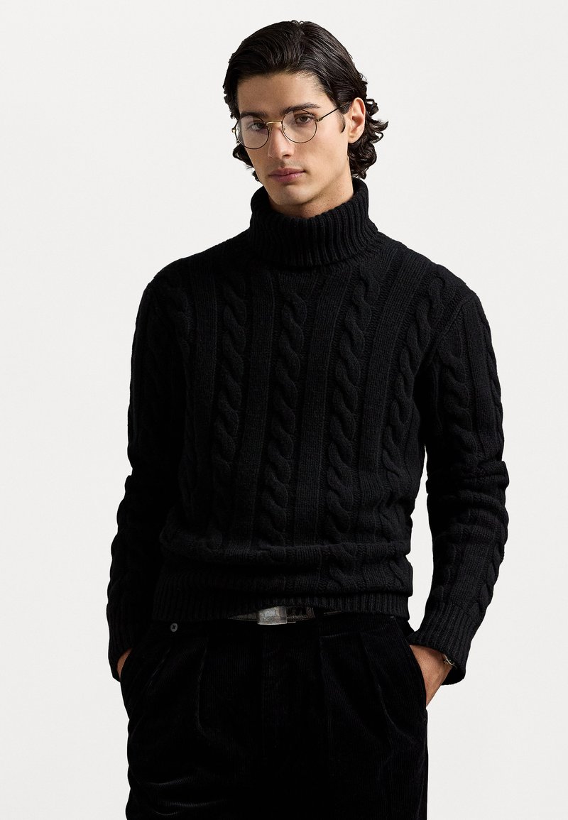 Melns kabela adījumā turtlenecks džemperis ar ribota apmala un piedurkņu galiem, ar teksturētu rakstu. Valkā ar tumšām biksēm, radot monohromu izskatu.