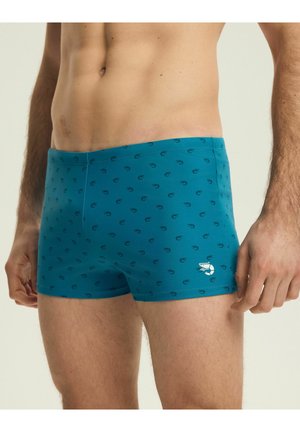 Mužský trup v tyrkysových plavkách s malými tmavými vzormi a bielym logom krevety na ľavom stehne.