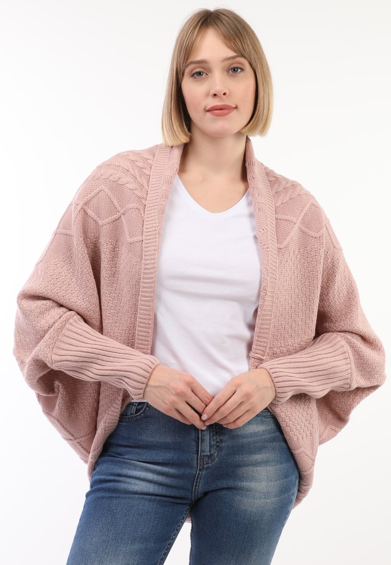 Cárdigan de punto rosa con frente abierto, patrones texturizados, mangas acanaladas y corte relajado, combinado con una camiseta blanca y unos vaqueros azules.