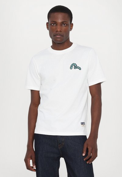 Camiseta blanca de algodón con mangas cortas, que presenta un diseño de ola bordado en verde en el lado izquierdo del pecho. Se combina con jeans azul oscuro.
