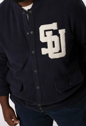 Mężczyzna noszący granatową kurtkę na guziki z białymi literami „SDU” na lewej piersi oraz kieszeniami z patkami na wysokości talii.