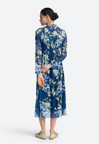 Cvetna midi obleka v modrih odtenkih z vzdržljivimi dolgimi rokavi, nabranim zgornjim delom in tekočo krilom. Kombinirana s svetlo obarvanimi, ravnimi čevlji.