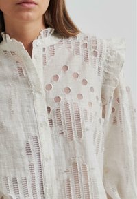 White linen shirt with cutout patterns and frayed edges. Features a ruffled collar and buttons down the front. Textured and lightweight.