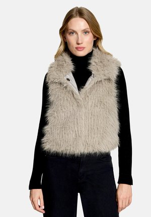 Donna che indossa un gilet in pelliccia sintetica beige chiaro sopra una maglia nera a maniche lunghe e pantaloni scuri, rivolta in avanti con espressione neutra.
