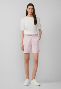 Maglione bianco a maniche corte abbinato a pantaloni corti rosa chiaro con risvolti, tasche frontali e una cintura intrecciata beige. Sneakers bianche completano l'outfit.