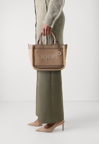 Borsa tote beige con dettagli in pelle color tan e una texture pelosa, con il logo "JOOP! JEANS" e un charm decorativo.