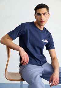 T-shirt en coton bleu marine à manches courtes et encolure ronde, arborant un logo blanc sur la poitrine. Le modèle est assis sur une chaise en bois.