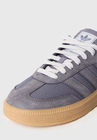 Szary sneaker wykonany z mieszaniny zamszu i siateczki, z białymi sznurowadłami, gumową podeszwą i trzema ciemnymi paskami po bokach.