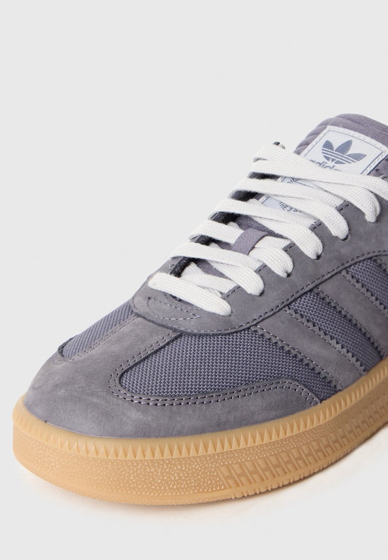 Grauer Sneaker mit einer Kombination aus Wildleder und Mesh-Materialien, ausgestattet mit weißen Schnürsenkeln, einer gummierten Außensohle und drei dunklen Streifen an der Seite.