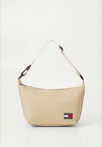 Bolso beige de hombro hecho de material liso, con una correa negra ajustable y un parche prominente con el logo "Tommy Jeans".