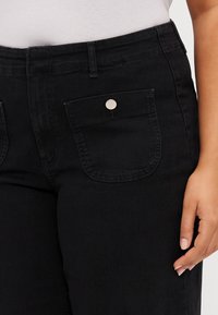 Svarta denimbyxor med hög midja, framfickor med silverdetaljer och en slät textur, en hand delvis synlig som vilar på tyget.