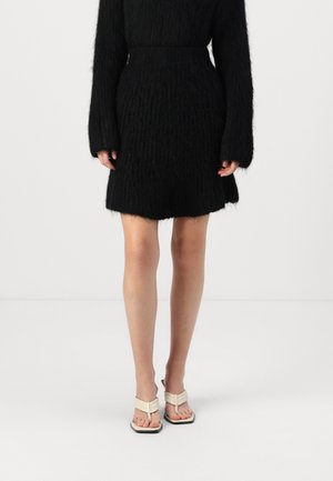 Svart strikket skjørt med en teksturert overflate, utsvingt kant, kombinert med lyse sandaler med et minimalistisk stroppesystem.