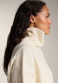 Pull à col roulé en tricot épais couleur crème, présentant des motifs de cable texturés et des détails côtelés, conçu avec une coupe ample et un col haut.