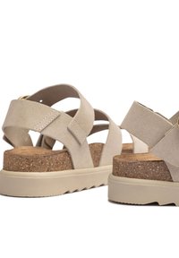 Beige suede sandals with crisscross straps, cork midsole, and thick rubber outsole featuring a textured pattern. Gold hardware accent detail.