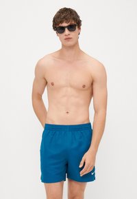 Kék úszónadrág rugalmas derékkal, a bal oldalon egy kis fehér Nike logóval. Az anyag könnyűnek és simának tűnik.