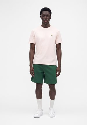 Mand stående iført lyserød t-shirt, grønne shorts, hvide sokker og hvide sneakers mod en ensfarvet hvid baggrund.
