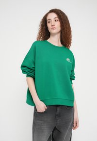 Πράσινο φαρδύ φούτερ με ριμπ μανσέτες και τελείωμα, με κεντητό σχέδιο στο στήθος. Φορεμένο με γκρι τζιν.